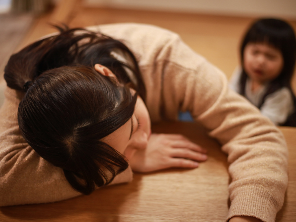 疲れた表情で机にうつ伏せる母親と、戸惑った様子の子ども