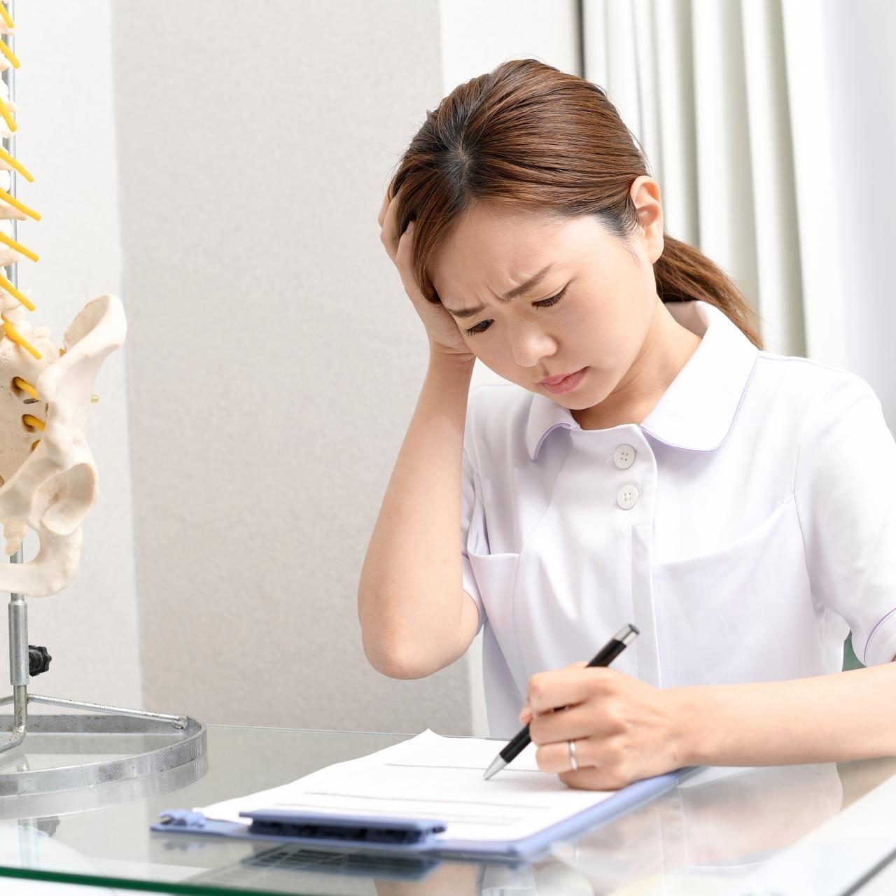 施術計画を立てながら頭を抱える女性整体師。経営や集客の課題に悩んでいる様子。