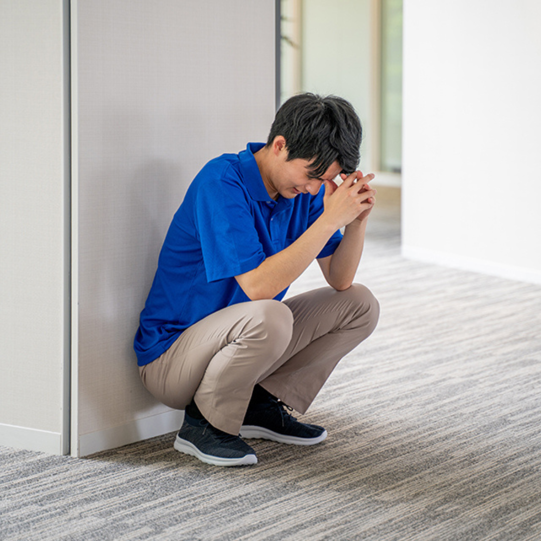 壁際でしゃがみ込み、不安な表情を見せる男性整体師。今後の経営や働き方に悩んでいる様子。