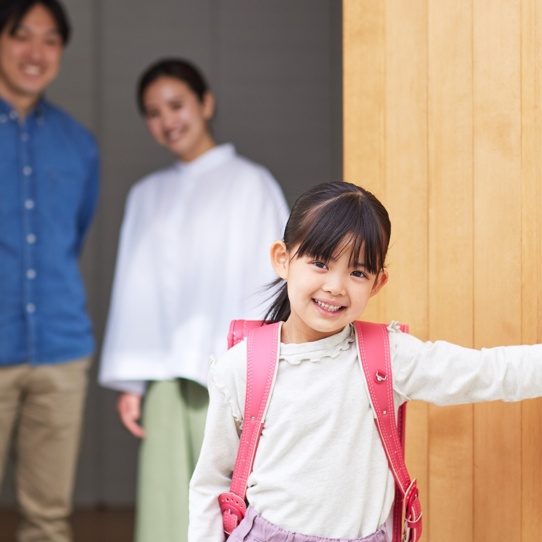 玄関で笑顔の女の子を見送りながら、後ろで親が穏やかに見守っている様子
