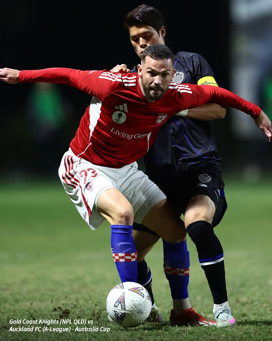 A male football player in a red kit dribbling the ball while being defended during a night match in Brisbane.