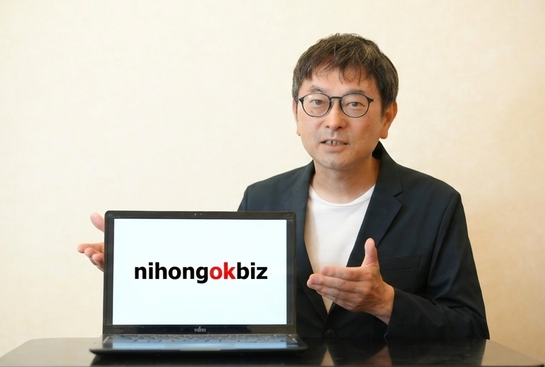 A man wearing a suit jacket gestures toward a laptop. The laptop screen shows the text "nihongok."