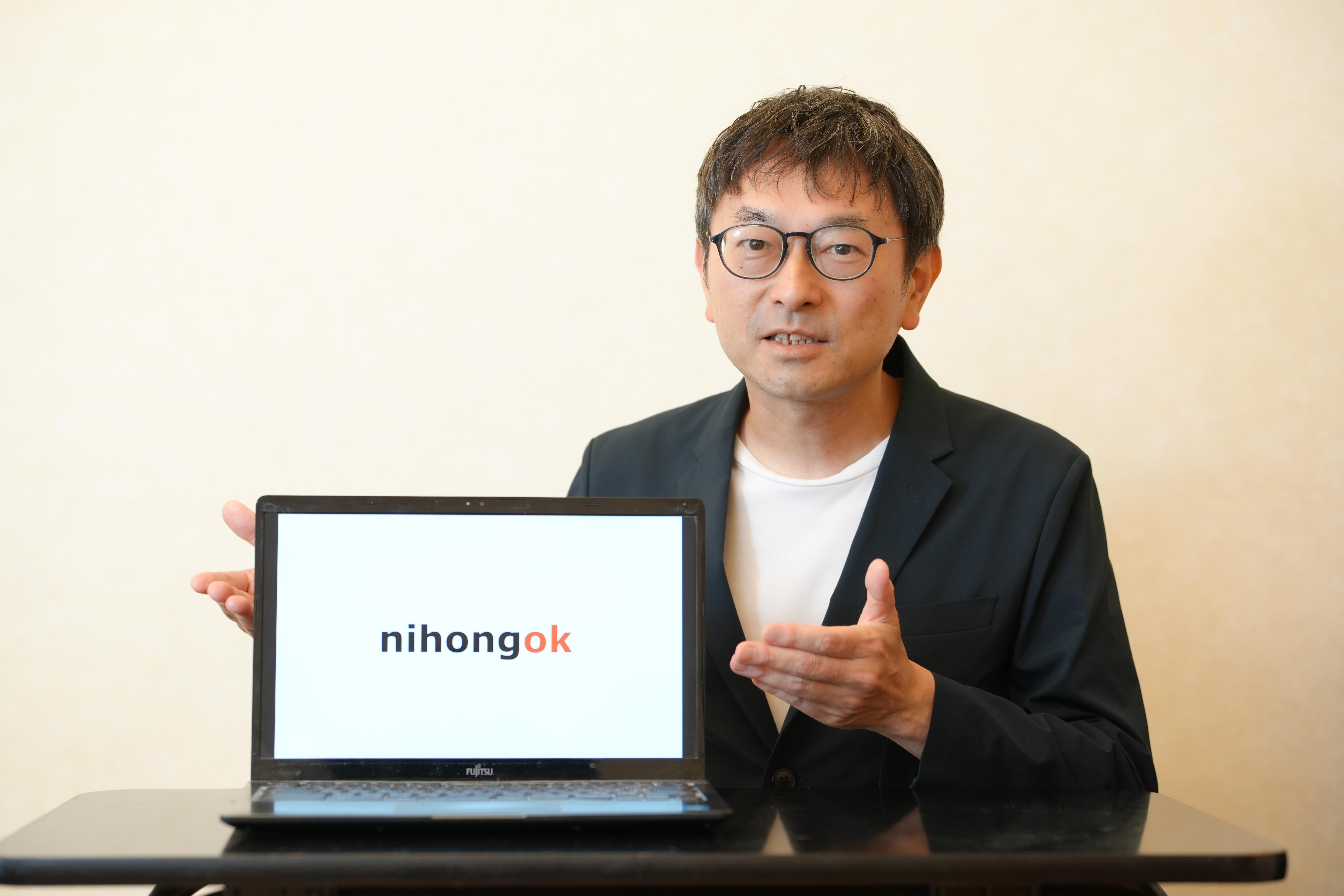 A man wearing a suit jacket gestures toward a laptop. The laptop screen shows the text "nihongok."