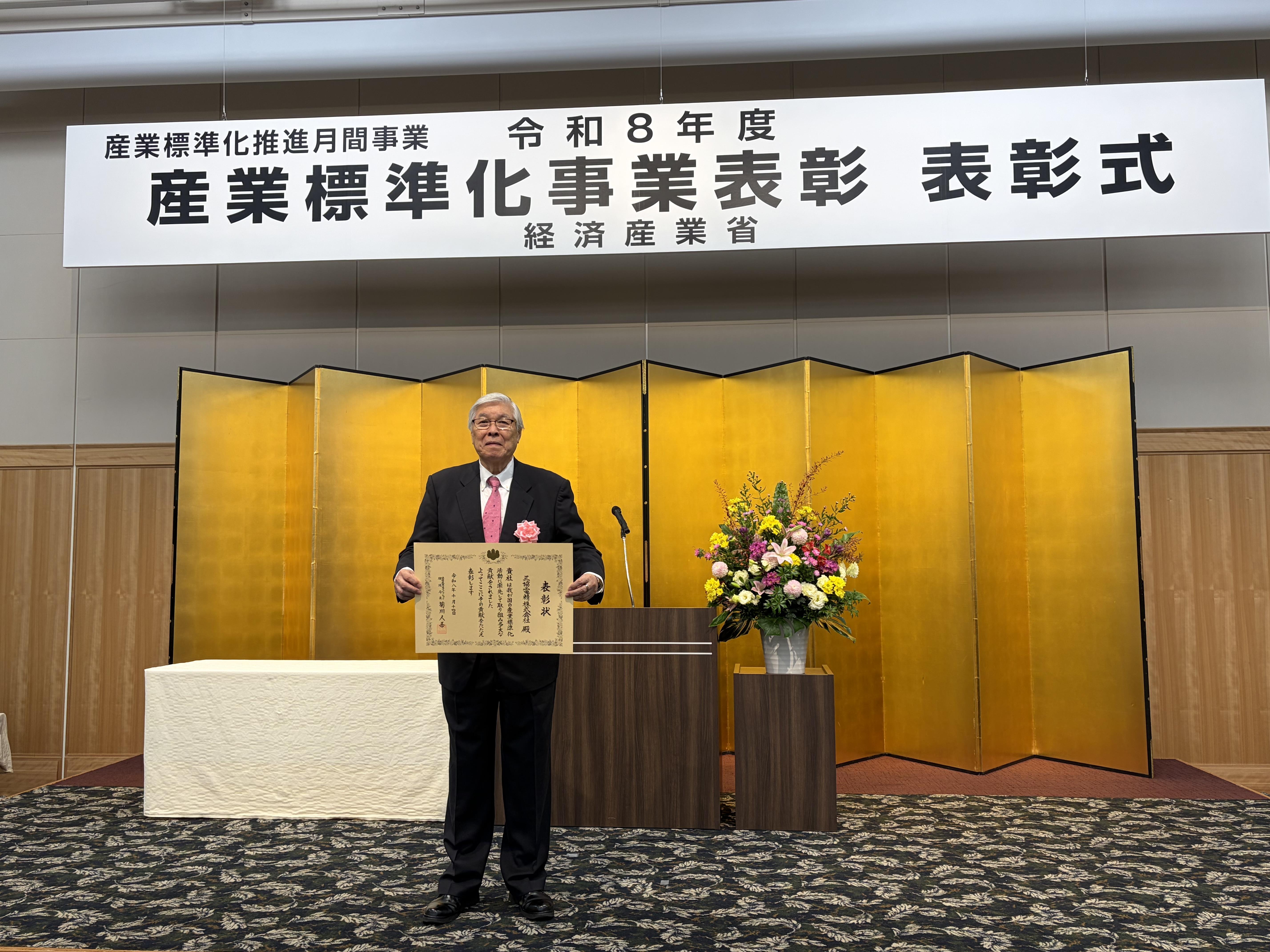 令和8年度産業標準化事業表彰の会場で、表彰状を手にする三協電精の受賞記念写真
