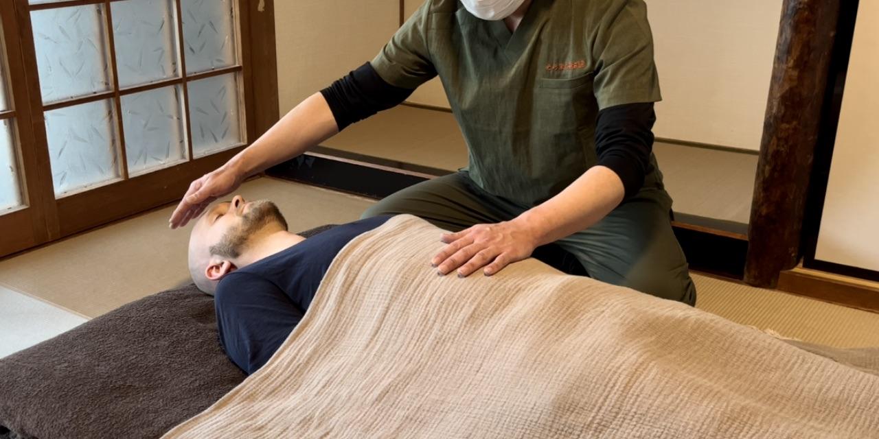A Reiki practitioner placing hands on the forehead and abdomen of a male guest from Norway lying on a futon, balancing the flow of Reiki energy.