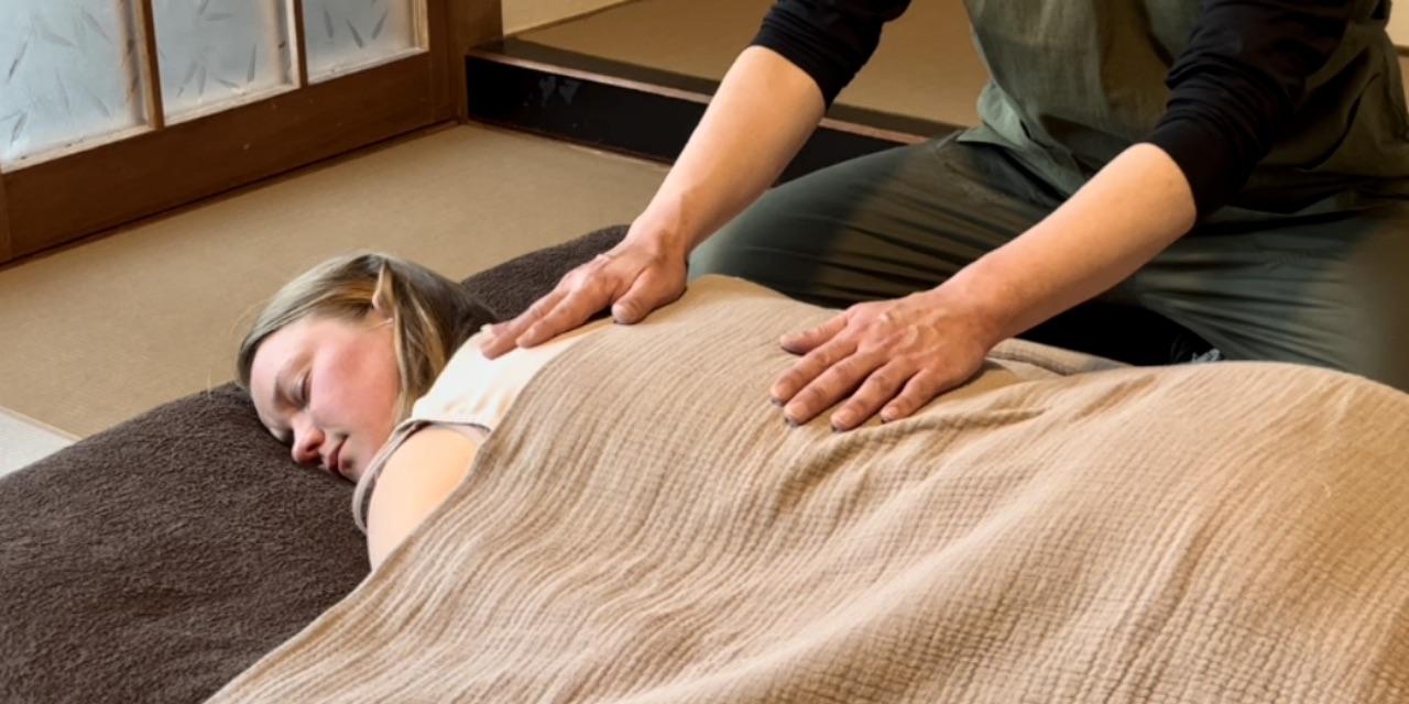 A Reiki practitioner placing both hands on the back of a female guest from Norway lying face down on a futon, encouraging deep breathing and relaxation.