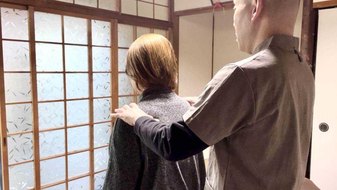 そらまめ整体院の院長が、自律神経の乱れに悩む女性客（ベッド端に座っている）の両肩に背後からそっと手を触れて、深呼吸を促しているところ。