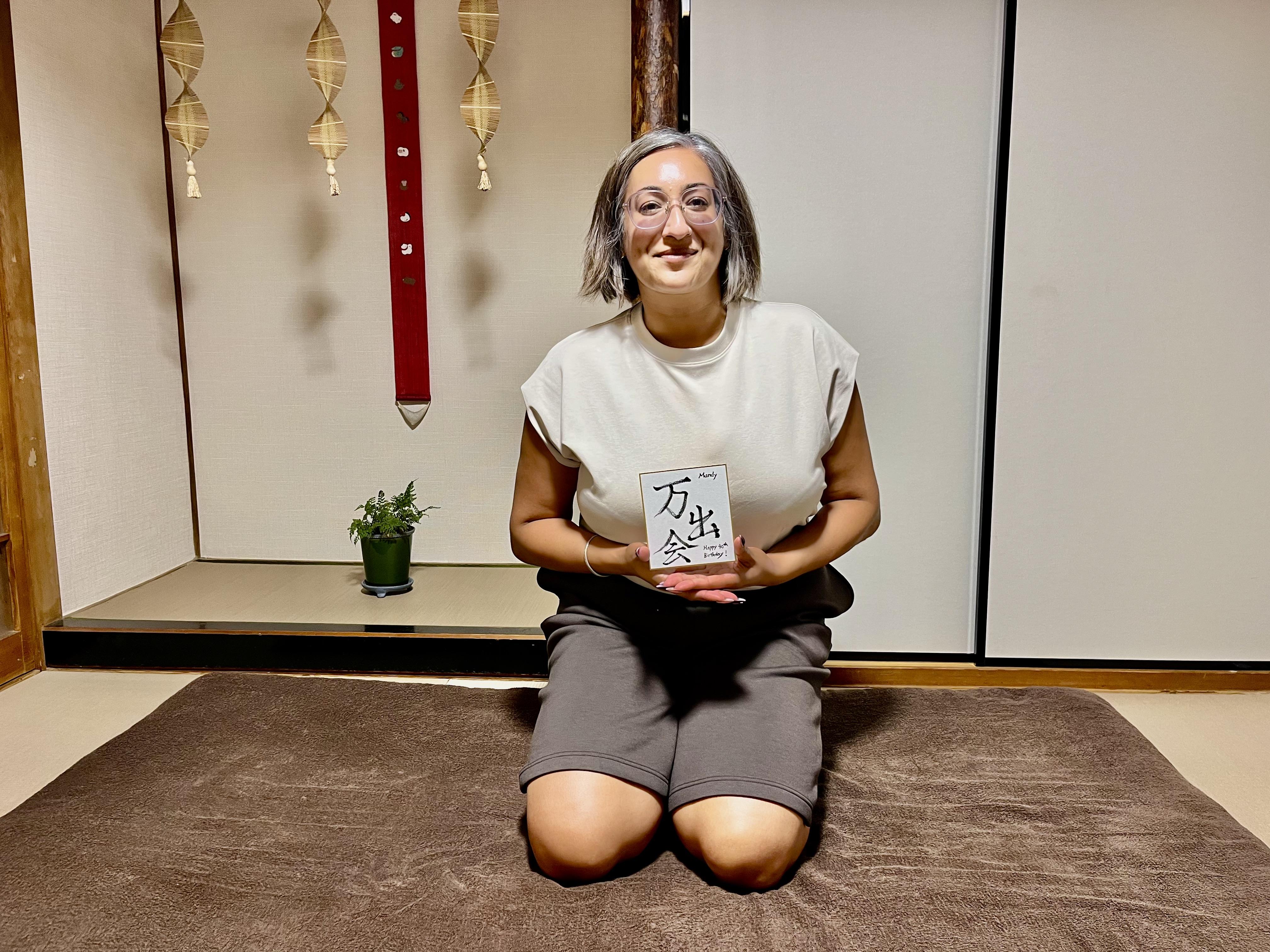 A commemorative photo of a smiling woman from UK, kneeling on a mat inside the traditional Japanese tatami room at Soramame Seitai Clinic, happily holding a personalized hand-written Kanji calligraphy gift that reads '万出会' (Mandy).