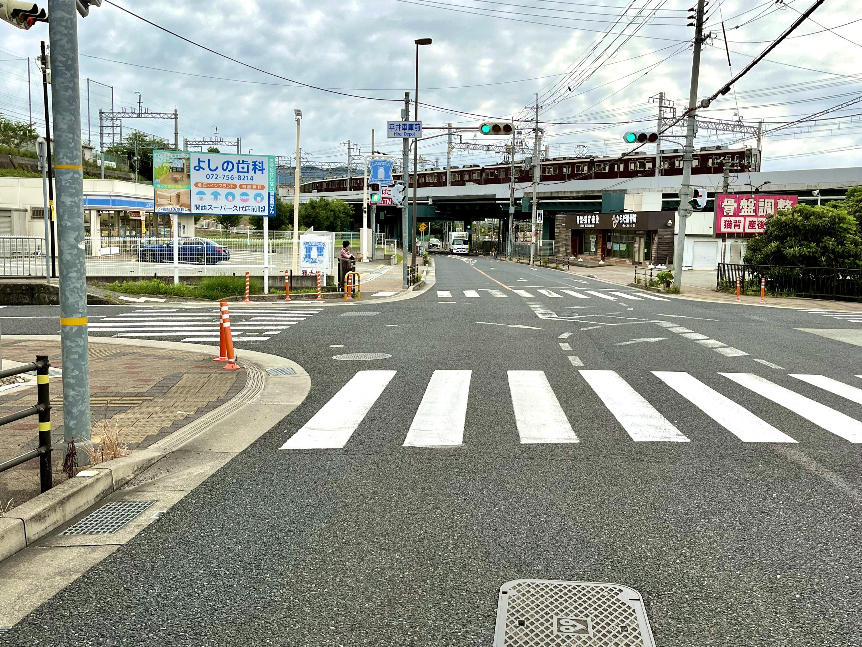 平井車庫前交差点の実際の写真｜山本から川西方向を臨む向きに撮られており、写真奥に阪急宝塚線平井車庫を走る阪急電車が見える。