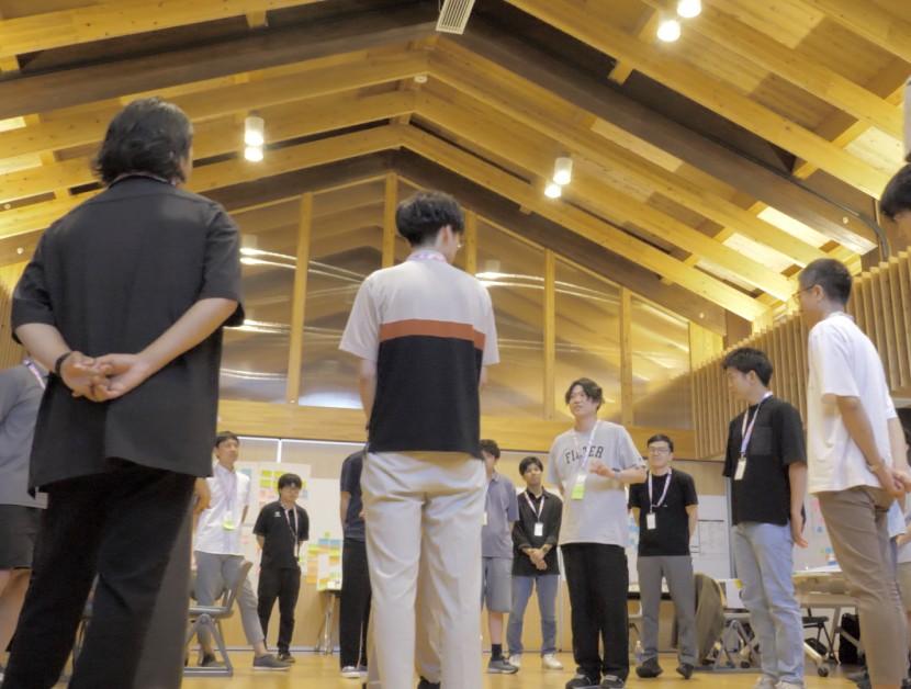 木造のホールで、参加者が輪になって向かい合い立つ様子
